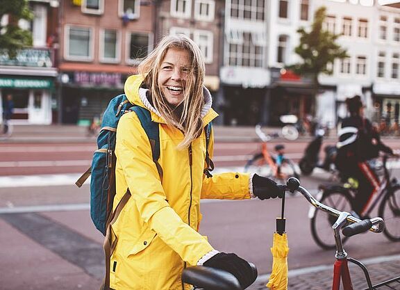 Symbolbild: Eine Frau in einer gelben Regenjacke steht lachend an ihrem Fahrrad, bereit loszufahren.