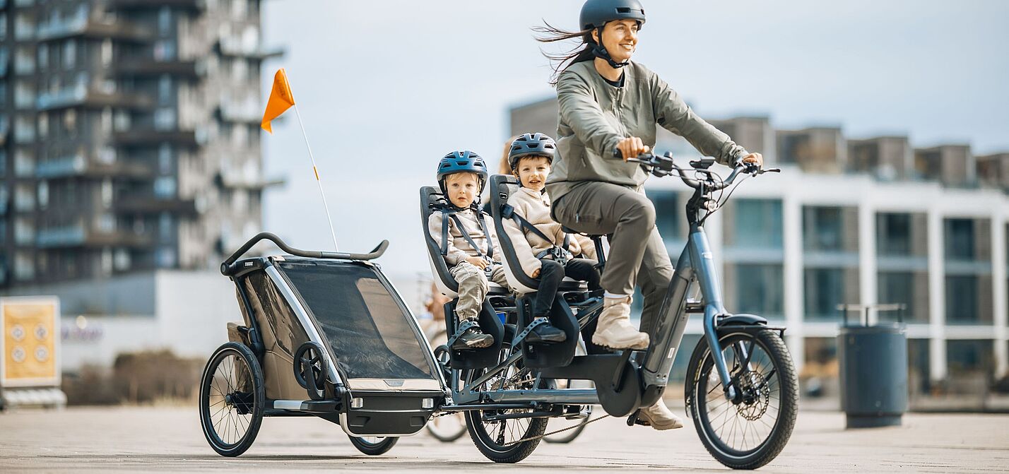 Familienlogistik: Longtail mit zwei Kindersitzen plus Anhänger Frau fährt ein Lastenrad Longtail, auf dem langen Gepäckträger hinten sitzen zwei Kinder in je einem Kindersitz. Das Fahrrad zieht zudem einen Kinderfahrradanhänger.